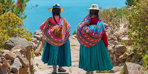Two indigenous Quechua women in traditional clothes walking down the path to the harbor of Isla Taquile (Taquile Island) with the Titicaca Lake in the background, Peru.